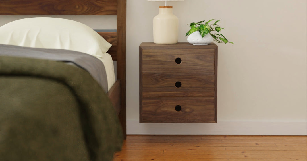 A wooden nightstand with three drawers, a ceramic lamp, and a small green plant sits next to a bed with a white pillow and dark green blanket on a wooden floor.