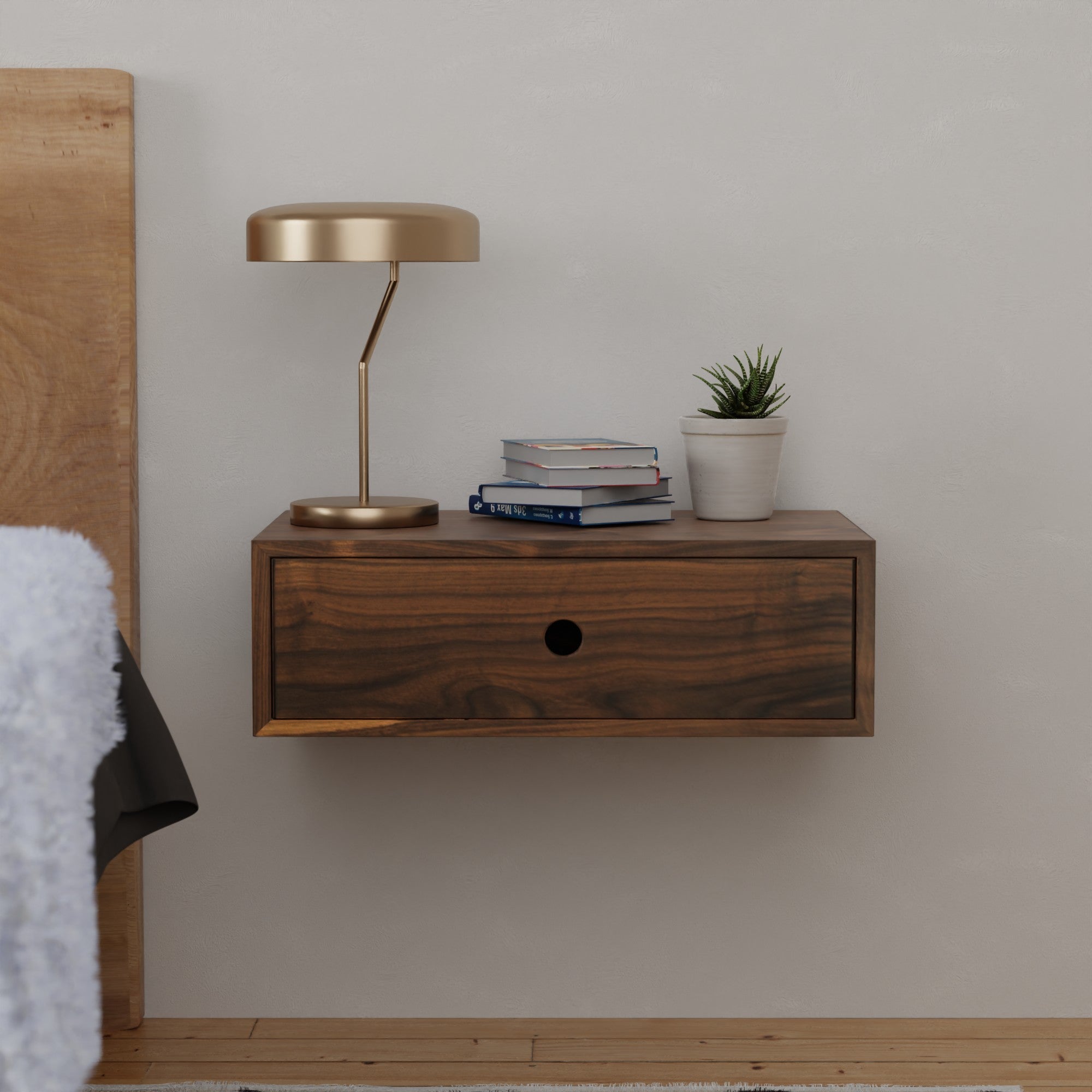 A modern bedroom nightstand with a single drawer, featuring a sleek gold lamp, stacked books, and a small potted plant. The nightstand is mounted on a light-colored wall beside a bed.