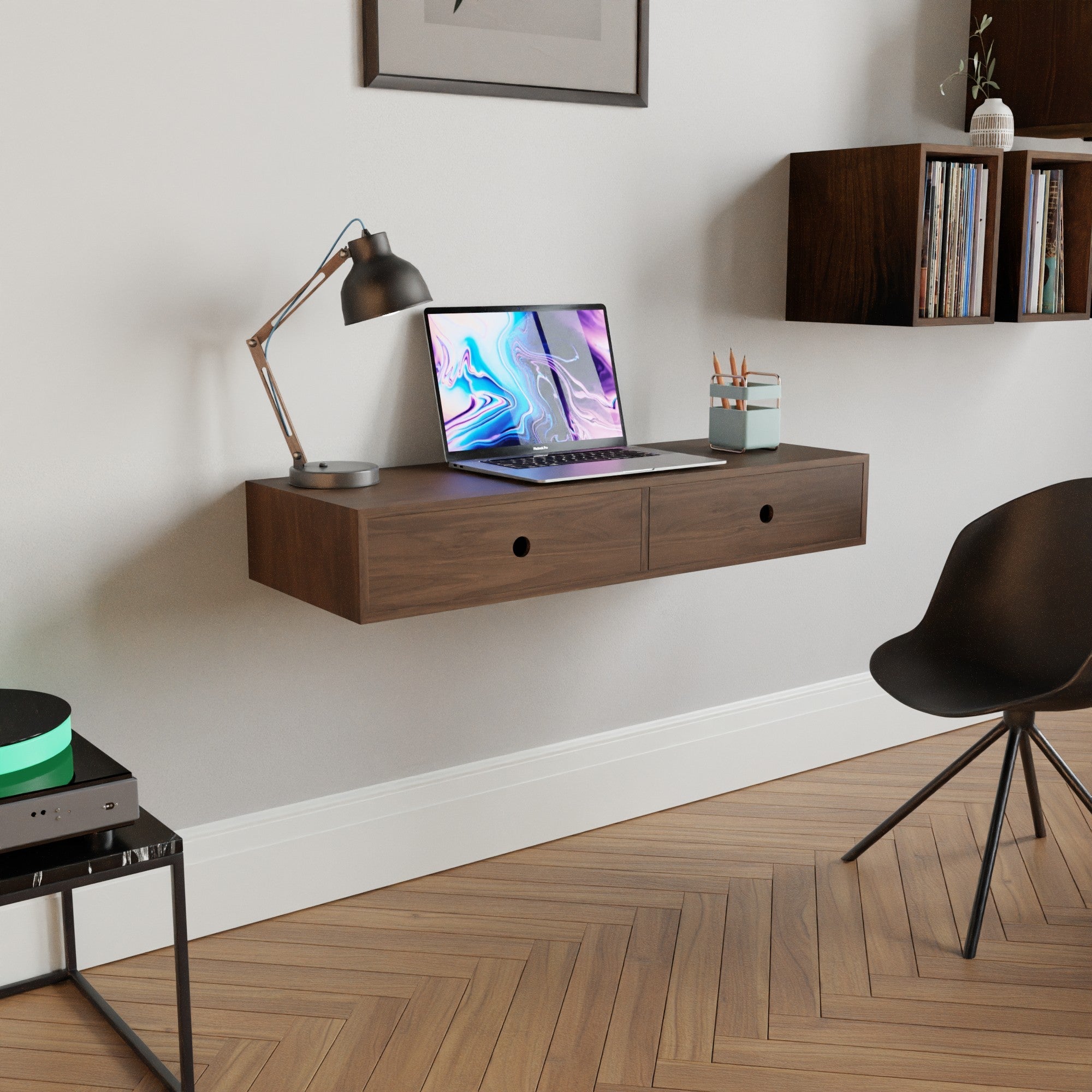 Minimalist workspace with a floating wooden desk, laptop, and lamp. A black chair sits nearby on a herringbone floor. Books are on a wall shelf, enhancing the modern aesthetic.