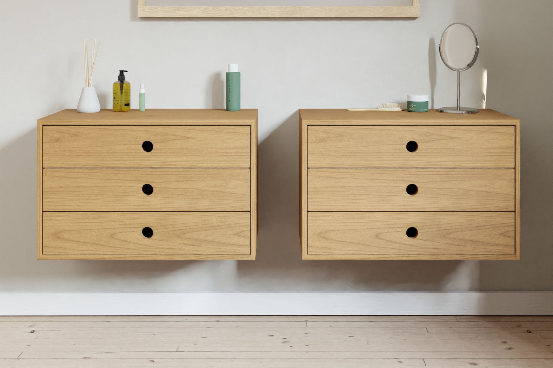 Minimalist interior design featuring two floating wooden cabinets against a light wall. Each cabinet has three drawers with black handles. On top are cosmetics, a mirror, and a reed diffuser. Light wood floor below.