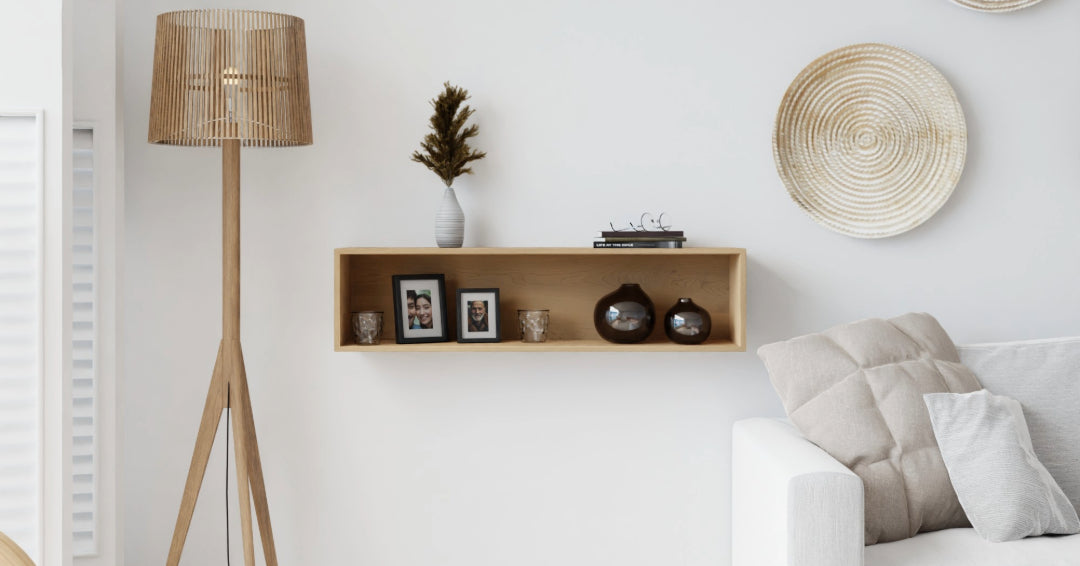 A minimalist living room with a white sofa, a wooden floor lamp, and a wall shelf displaying framed photos, vases, and a small plant. Two decorative woven plates hang on the wall. Neutral color scheme.