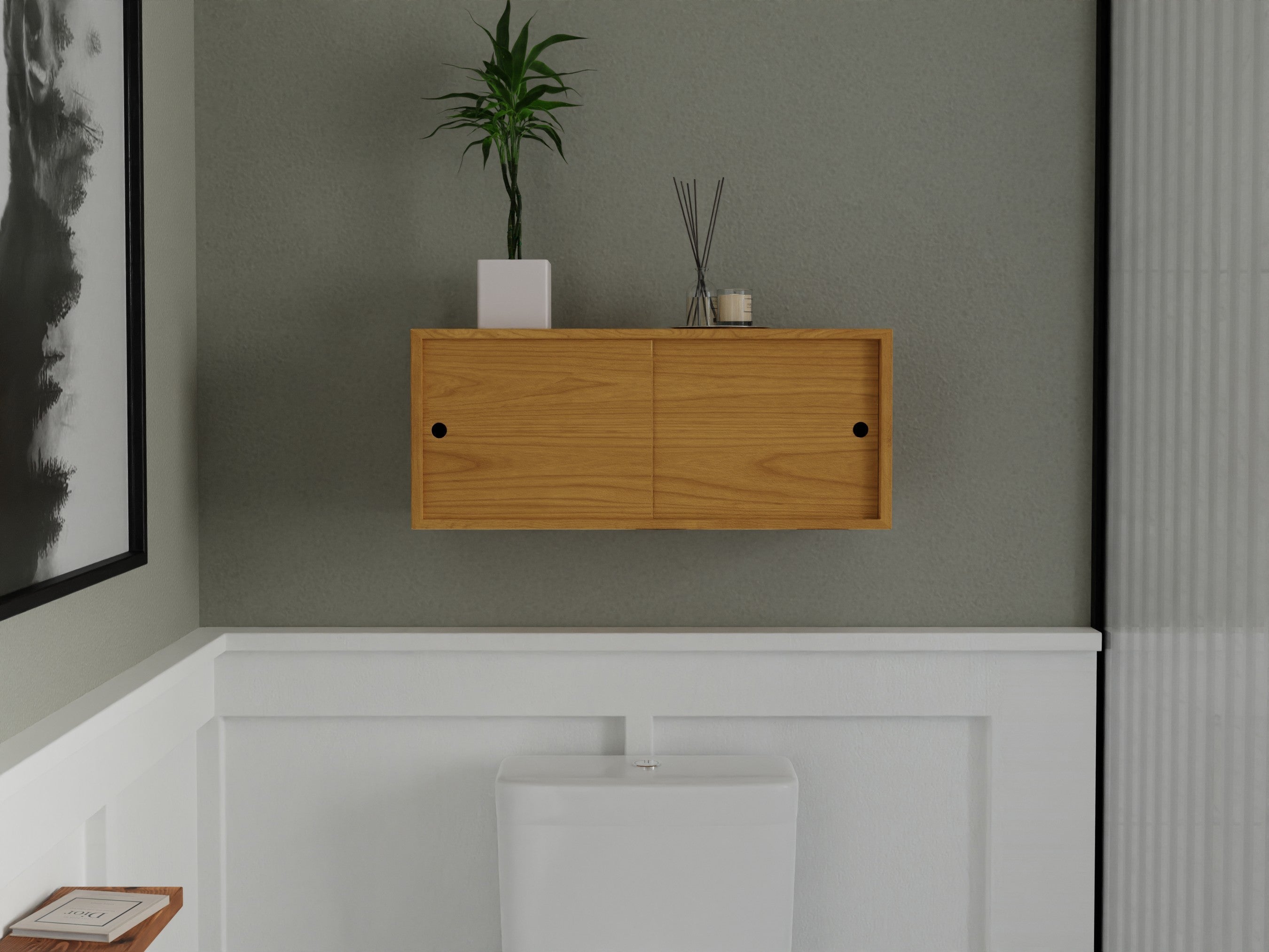 A minimal bathroom with a white toilet, wooden wall cabinet, and potted plant on top. The walls are a light gray-green, and there is a black-and-white abstract painting on the left.