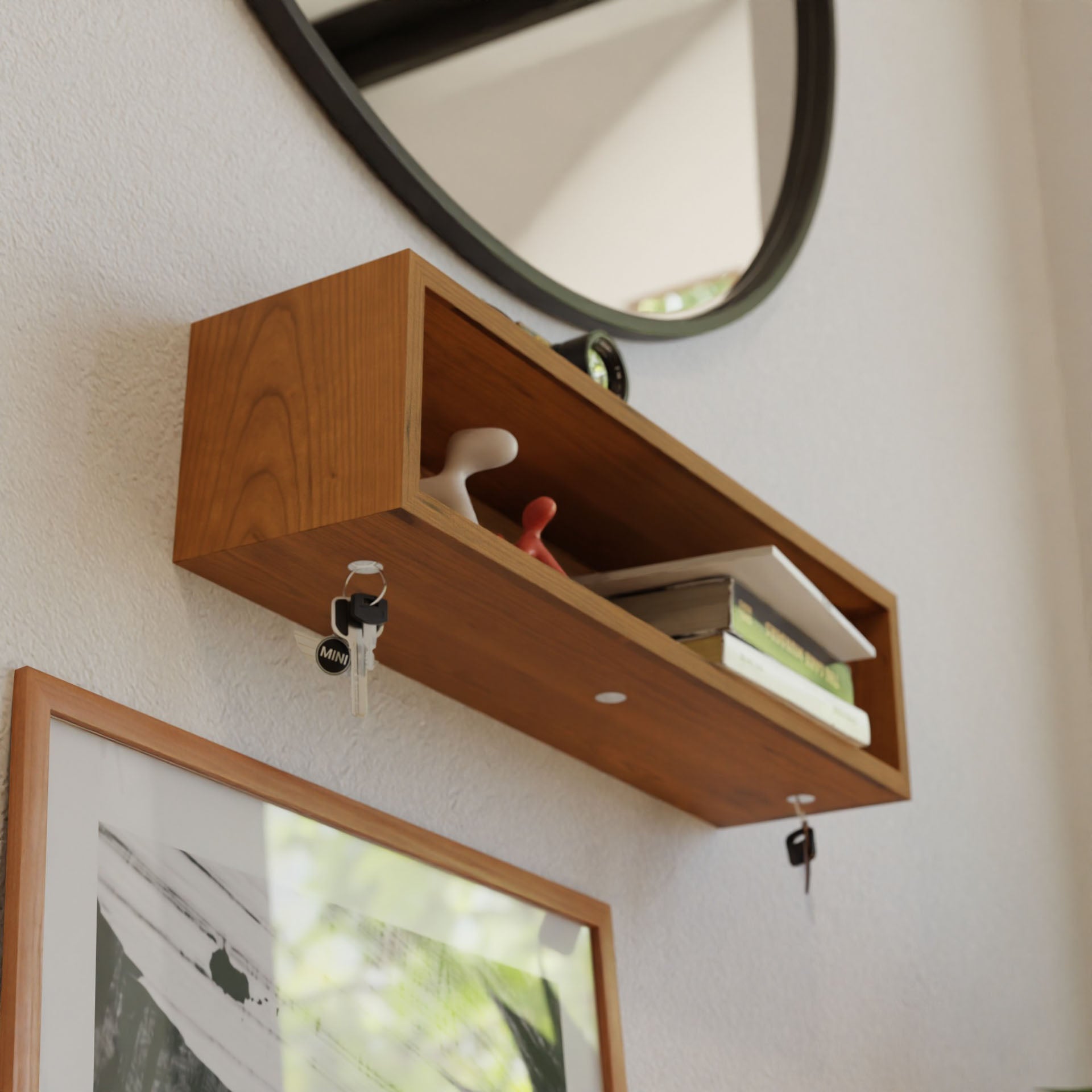 A handmade, wooden wall-mounted shelf holds a few books and decorative items. Below the shelf, two keys hang from hooks. A round mirror is mounted above, and a framed picture partially visible below the shelf, all crafted with care in the USA.