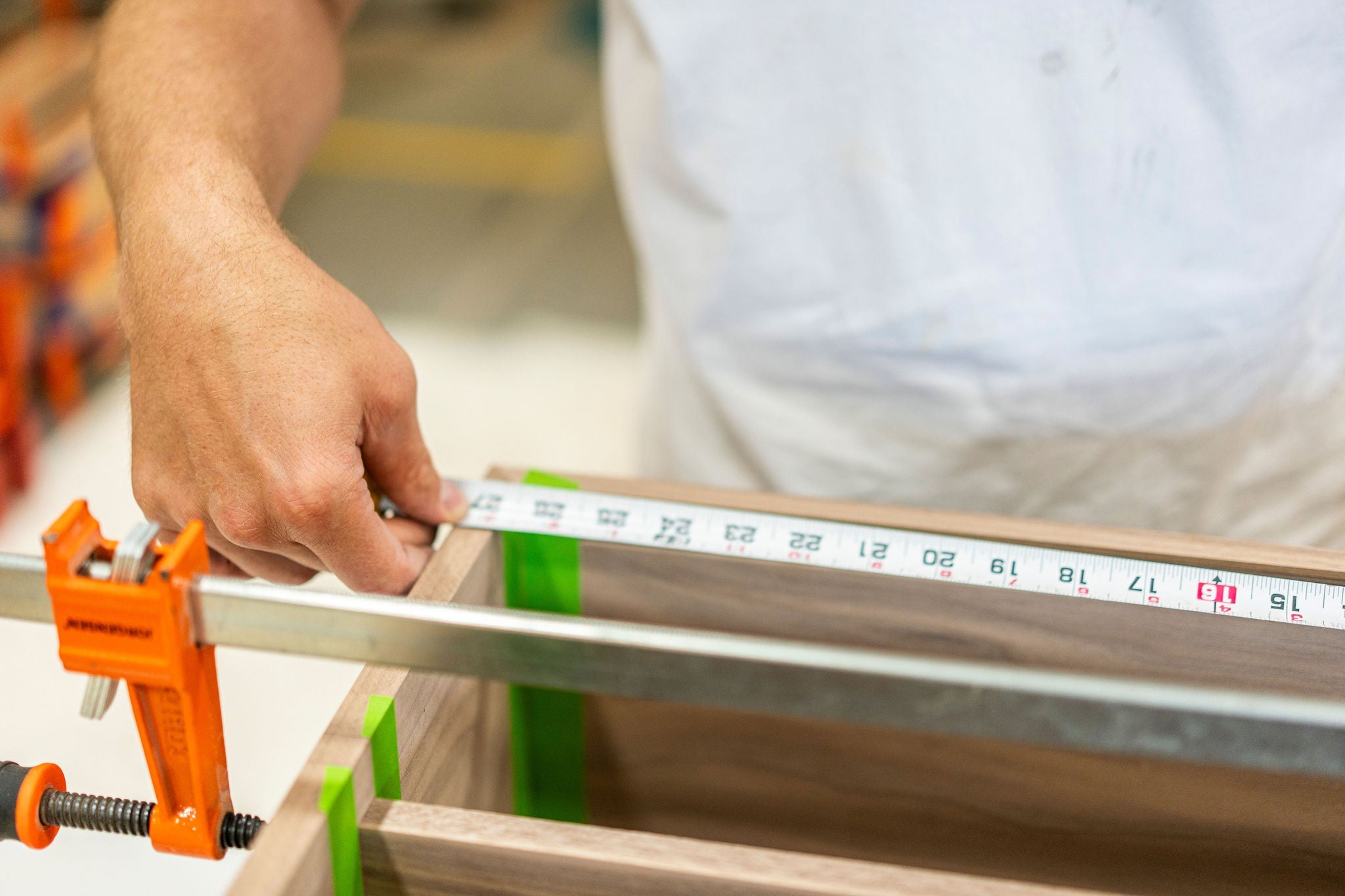 A person from Krovel Furniture Co., known for their premium hardwood creations, uses a tape measure to check the dimensions of a wooden panel secured with an orange clamp. The focus is on the hand and measuring tape, with the person wearing a crisp white shirt. Made in USA excellence at its best.