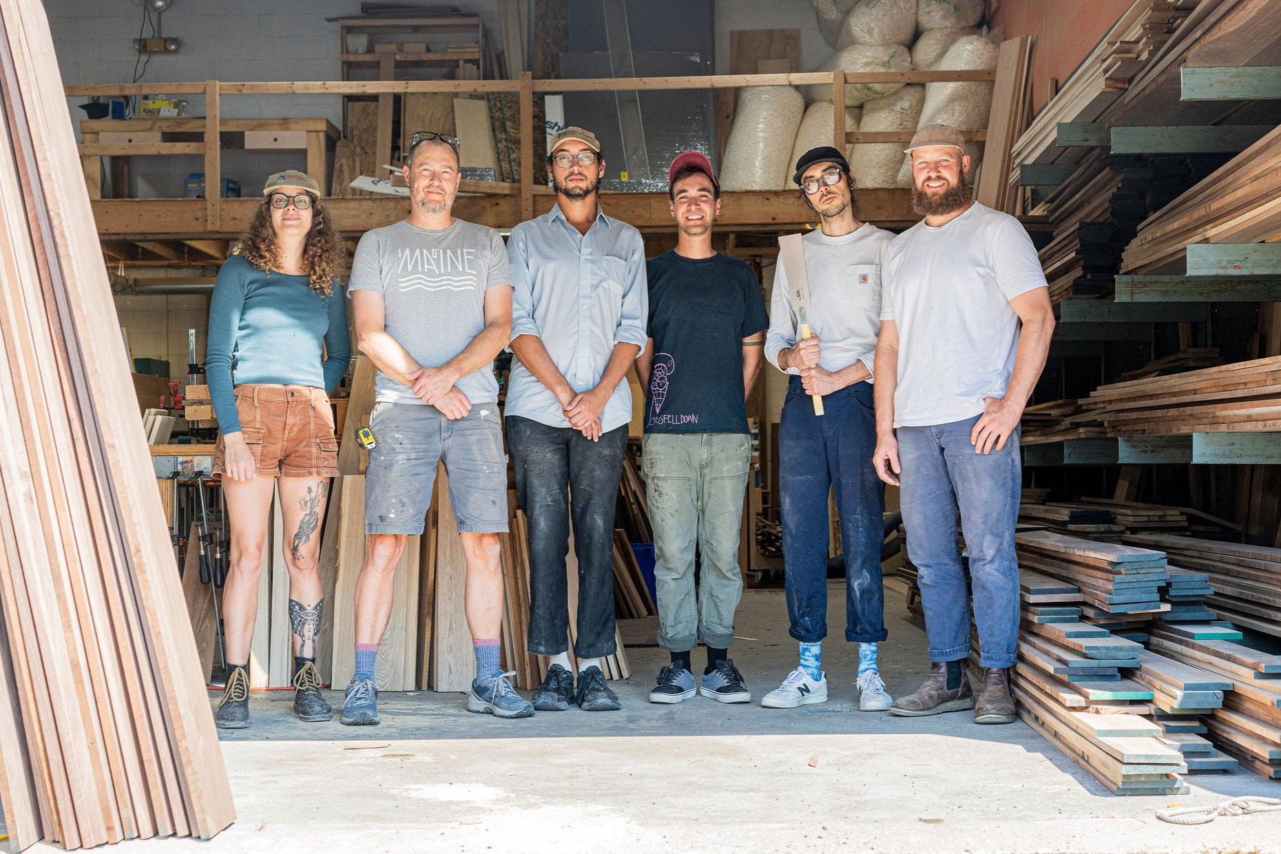 A group of six people, four men and two women, stand inside a woodworking workshop. Surrounded by stacks of hardwood planks and tools, they smile warmly amidst shelves filled with handmade creations. Dressed casually, their pride in crafting pieces made in the USA shines through.