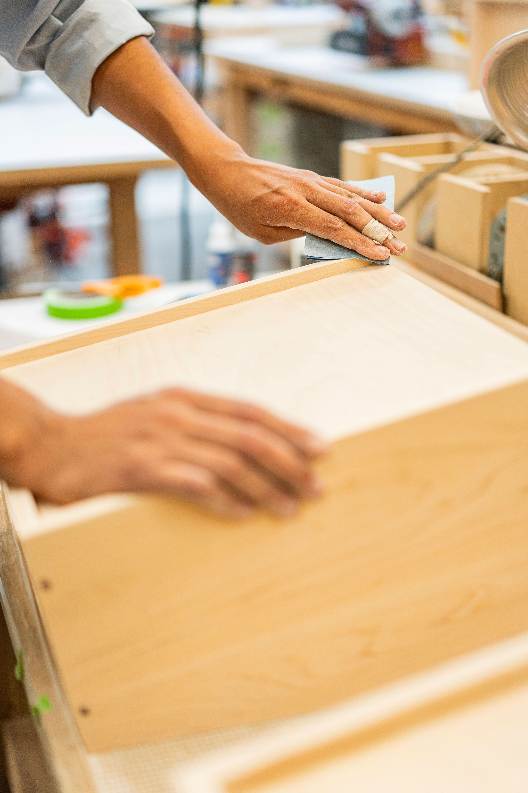 In a bustling workshop, a craftsman from Krovel Furniture Co. is meticulously sanding the edge of a handmade wooden box. Using sandpaper to perfect the finish, their skilled hands glide over the surface, surrounded by tools and materials that highlight the artistry of Made in USA craftsmanship.