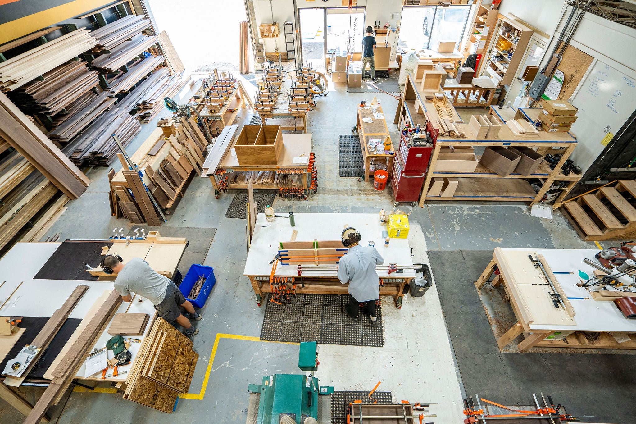 Aerial view of a Krovel Furniture Co. woodworking workshop with two people crafting projects. Various hardwood materials and tools are organized on shelves and workbenches. Natural light streams in from large windows, illuminating this proud Made in USA space.