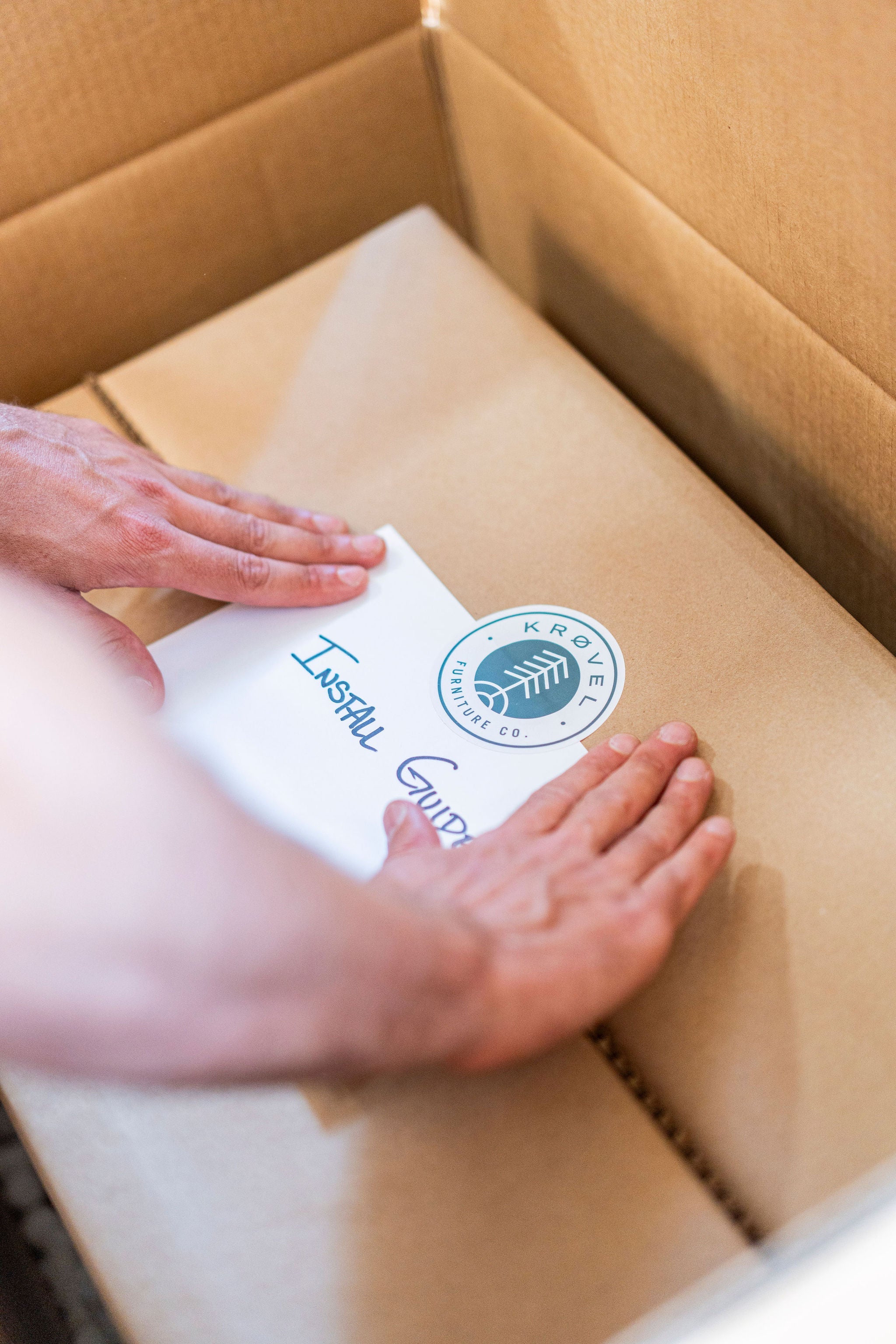 A person is placing an installation guide with a "KROVEL" logo on a cardboard box. The guide, featuring a green leaf within a circle, signifies the attention to detail by Krovel Furniture Co., renowned for its handmade hardwood pieces. The scene suggests packaging or preparing an order for shipping.