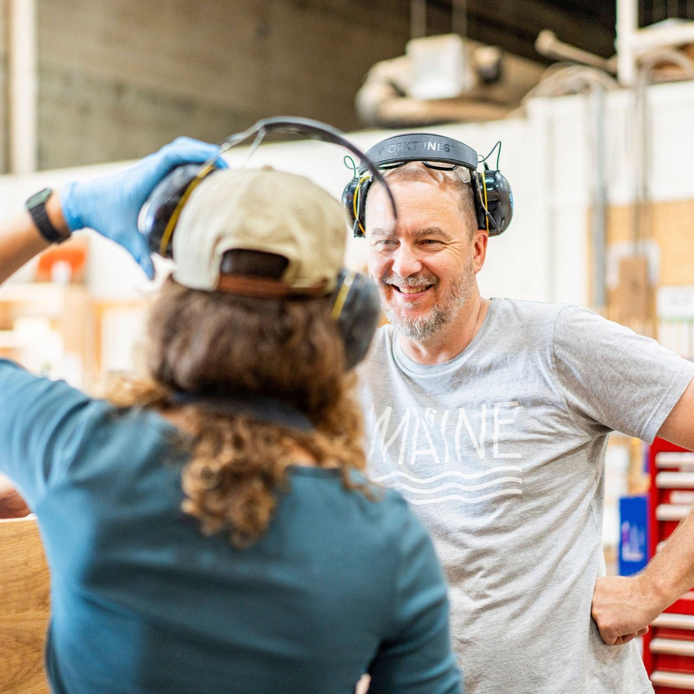 In a bustling workshop, two individuals don hearing protection. One wears a gray shirt marked "Maine" and smiles warmly, while the other sports a cap with long hair flowing behind. Surrounded by tools and equipment, they craft exquisite pieces for Krovel Furniture Co., specializing in handmade hardwood designs.