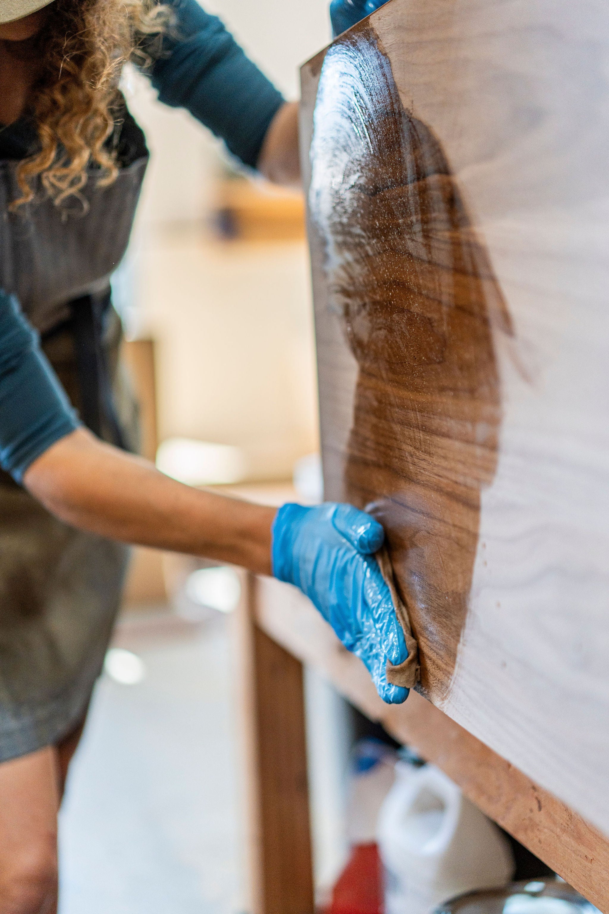 A person wearing blue gloves is carefully applying varnish to a handmade hardwood surface in a workshop. The wood, crafted in the USA, boasts a shiny finish where the varnish has been applied. The person is dressed in a work apron.