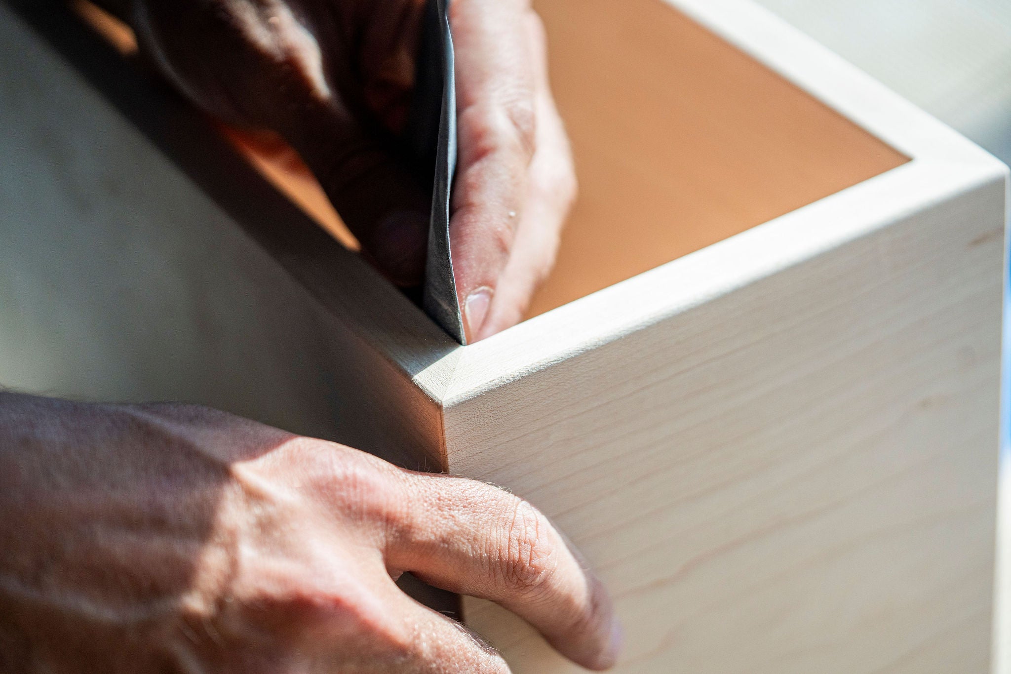 Close-up of hands smoothing the edge of a wooden box with sandpaper. The sunlight highlights the fine grain, emphasizing Krovel Furniture Co.'s craftsmanship in the handmade woodworking process, proudly made in the USA.