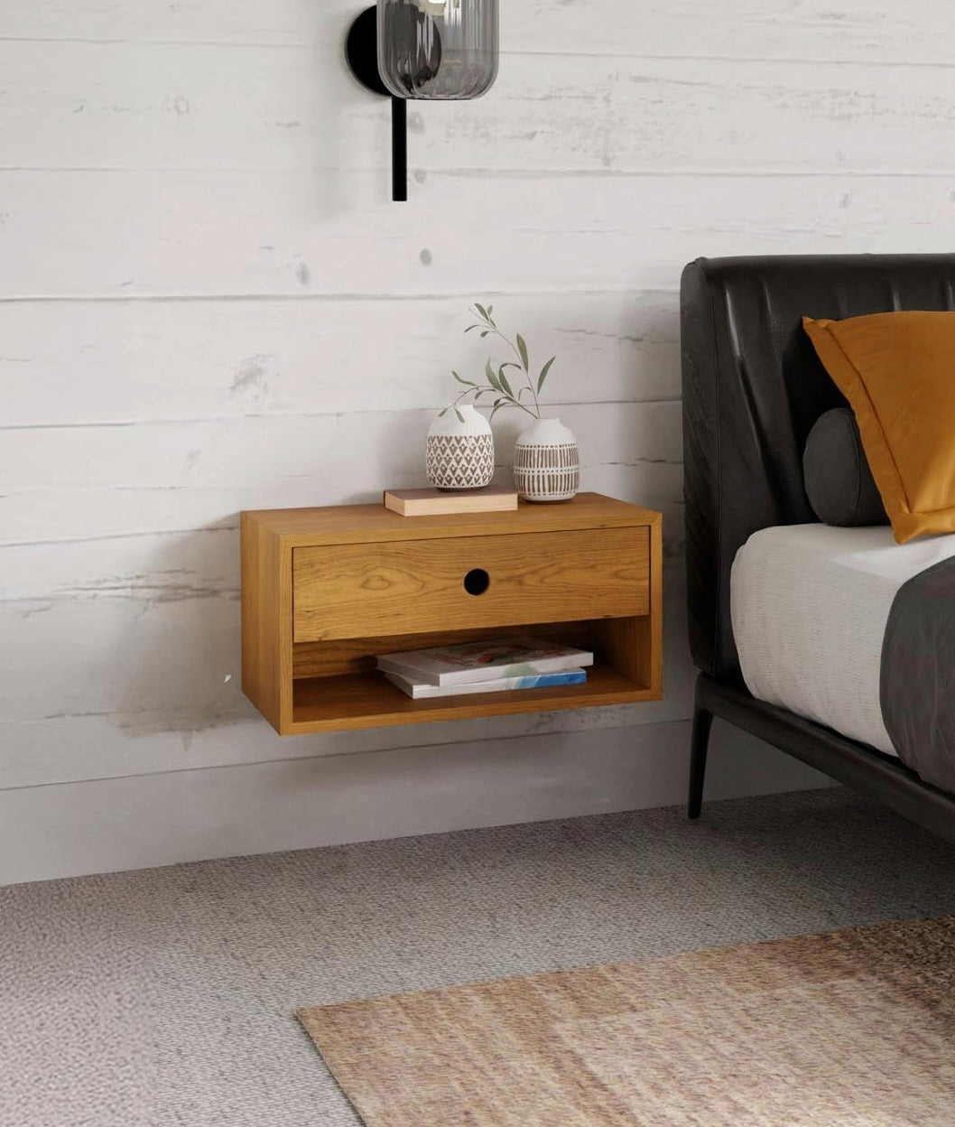 A modern bedroom features a floating wooden nightstand by Krovel Furniture Co. on a white shiplap wall. The nightstand, made in the USA, holds a small plant and stacked books. Part of a bed with a gray headboard and mustard yellow pillow is visible, with a sconce light mounted above.