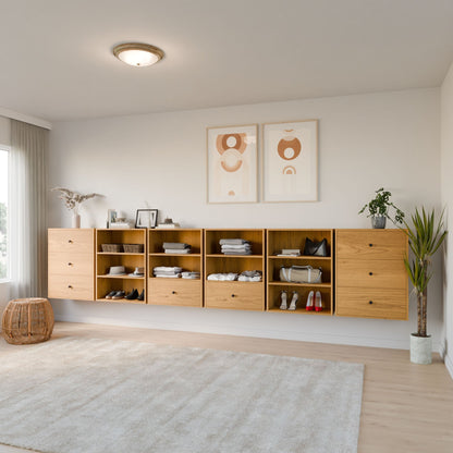 A minimalistic room showcases Krovel Furniture Co.'s Closet Drawers and Shelves in Cherry, organized with folded clothes, baskets, and shoes. Two abstract artworks adorn the wall above. A potted plant and wicker stool are on the floor, while a large window invites natural light in. 