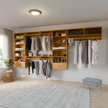 A minimalist closet displays Closet Drawers and Shelves in Cherry by Krovel Furniture Co., featuring wooden hanging rods on a white wall. Clothes, shoes, and storage boxes are neatly organized. A gray ottoman and a plant in a wicker basket enhance the light wood floor, bathed in natural light. 