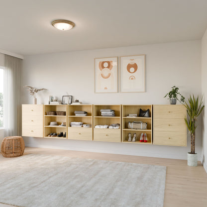 A minimalist bedroom showcases Krovel Furniture Co.'s Closet Drawers and Shelves in Maple, displaying clothes, shoes, and decor. Two abstract art pieces hang above. A large window with sheer curtains bathes the room in light. A wicker basket and potted plant complete the look. 