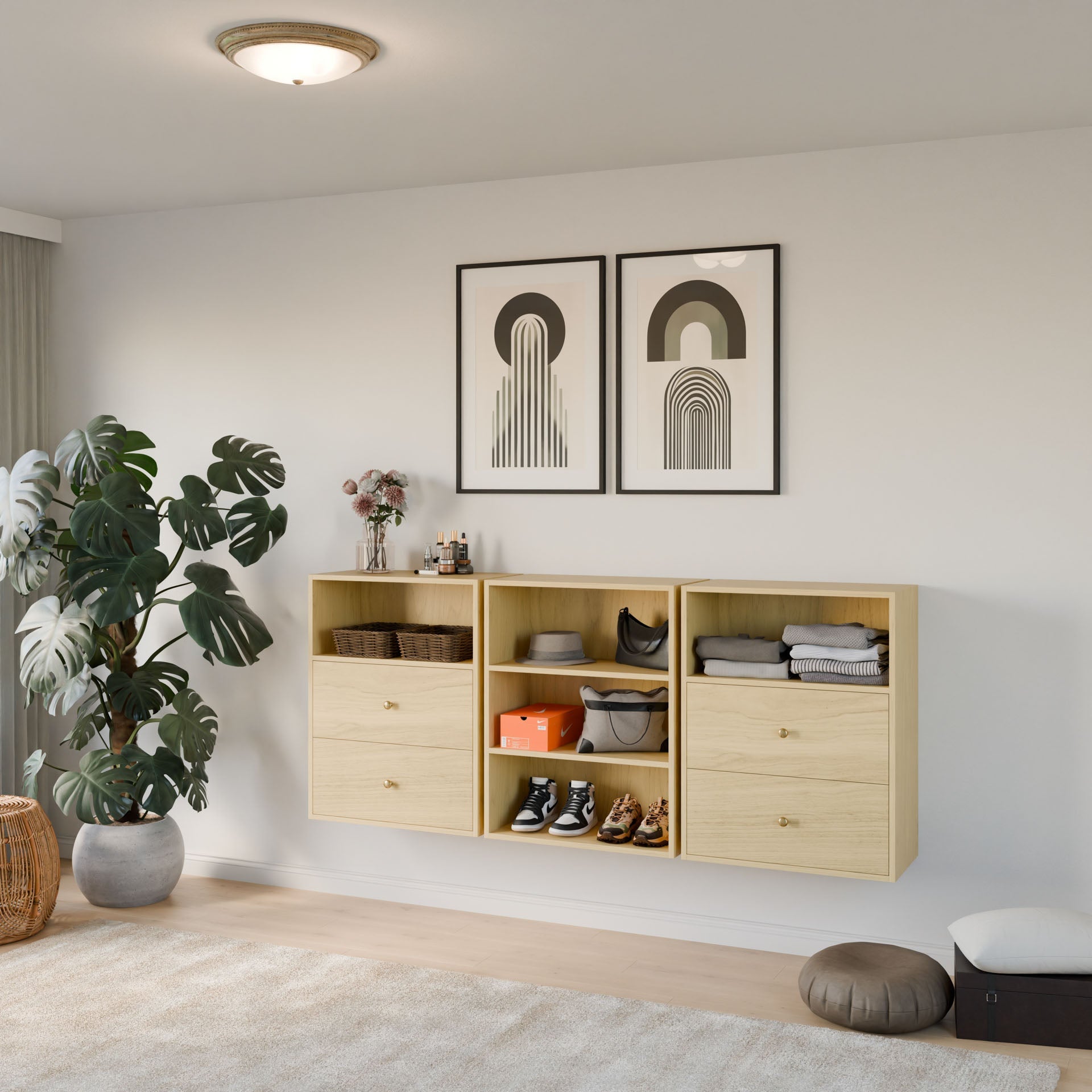 In a modern room by Krovel Furniture Co., the Closet Drawers and Shelves in Maple hold shoes, hats, and decor items. Two abstract art pieces are displayed above, while a large plant and brown pouffe sit on the carpeted floor, complemented by a round ceiling light. 