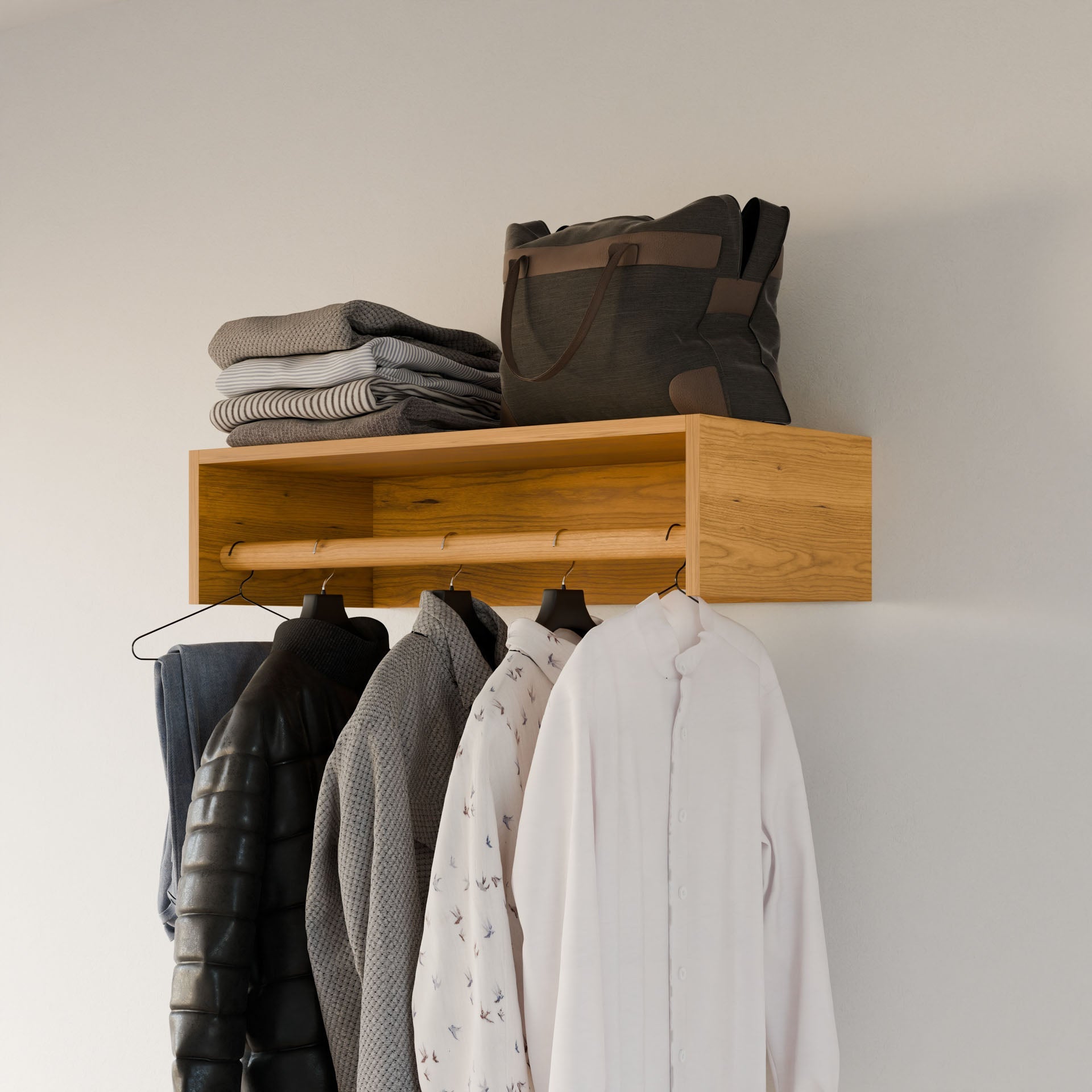 The Closet Hanger in Cherry by Krovel Furniture Co., a wooden, wall-mounted clothes rack handmade in Portland, Maine from premium-grade plywood, holds a black jacket, two gray sweaters, and a white shirt on hangers. Gray clothes and a gray tote bag rest neatly folded on top against a simple white wall.