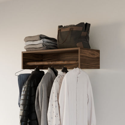 The hardwood veneer wall shelf displays folded sweaters and a dark bag. Below, a Krovel Furniture Co. Closet Hanger in Walnut showcases garments: a white shirt, patterned shirt, grey sweater, and padded jacket. The backdrop is light-colored.