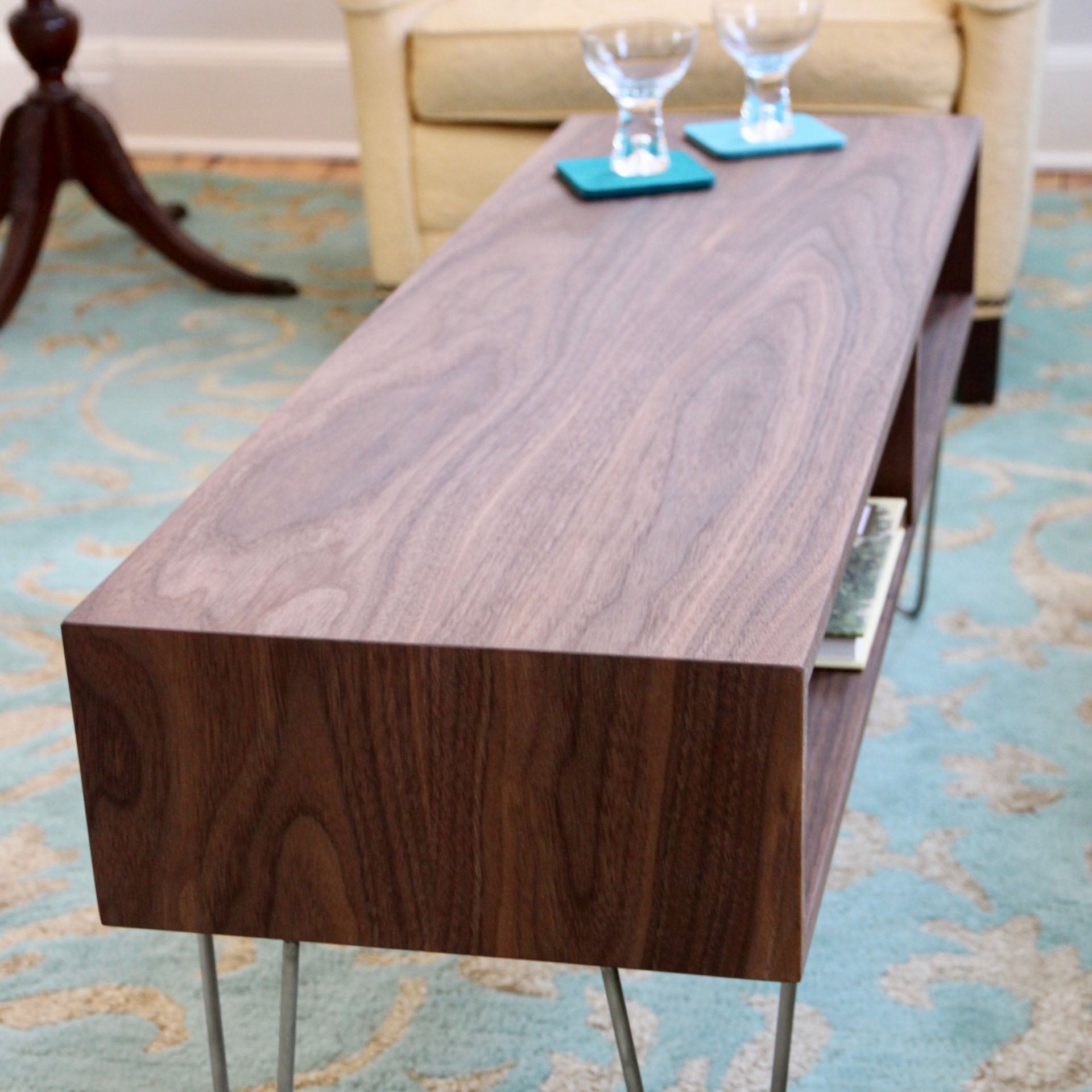 The Krovel Coffee Table in Walnut, featuring a mid-century modern design with a natural finish and stainless steel hairpin legs, serves as the perfect centerpiece. Two glasses on coasters are placed on top, while a book peeks out from the shelf underneath. This elegant black walnut piece is beautifully set on a patterned blue rug.