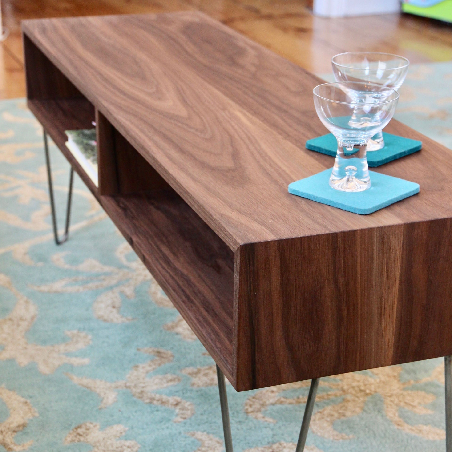 A mid-century modern coffee table from Krovel, named the Coffee Table in Walnut, features sleek stainless steel hairpin legs and holds two glass goblets on blue coasters, all resting on a patterned blue rug in the room.