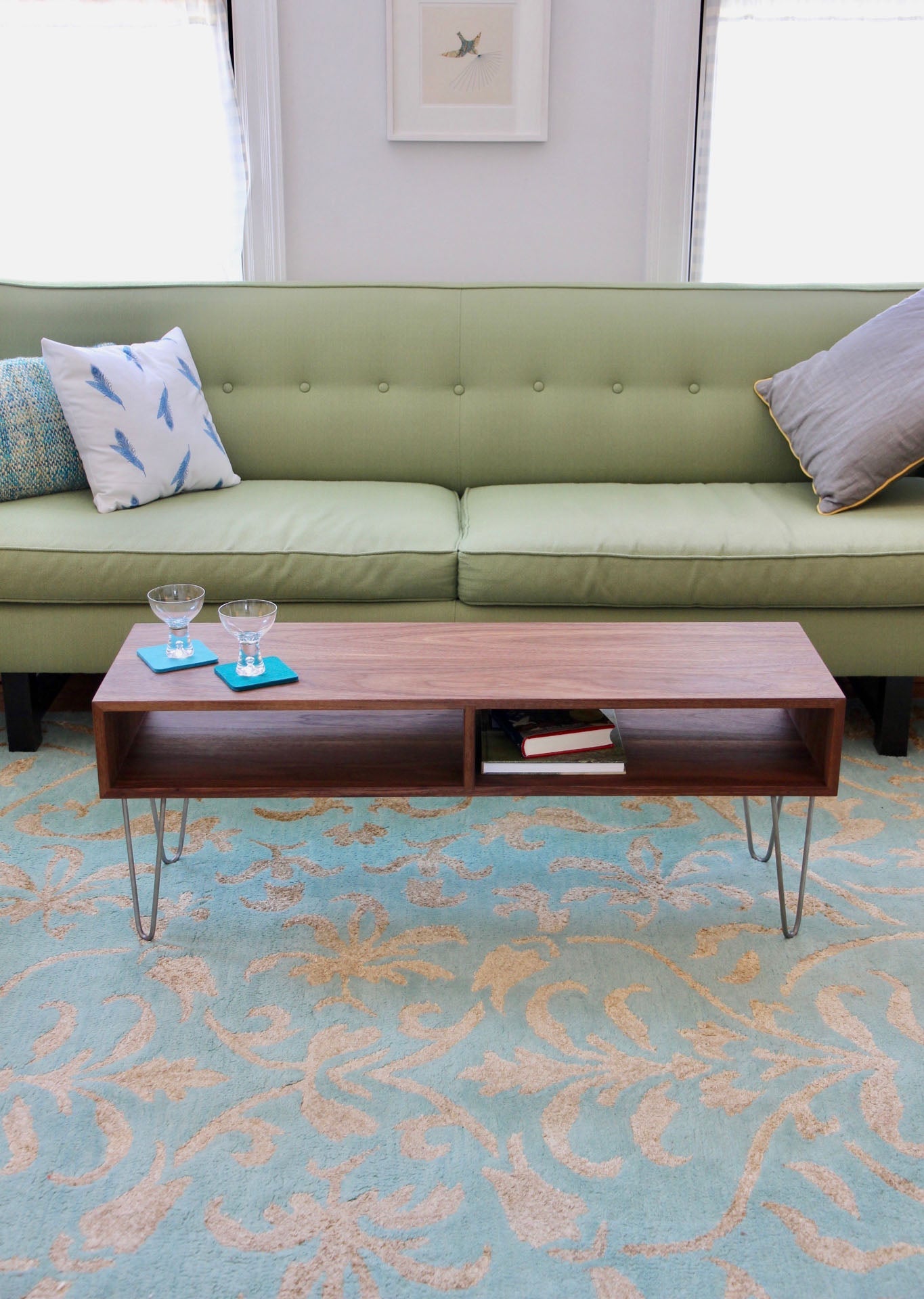 A contemporary living room showcases a light green sofa adorned with colorful pillows. A "Coffee Table in Walnut" by Krovel, featuring stainless steel hairpin legs, supports two small glasses. The floor is adorned with a patterned blue and beige rug, while a framed picture is displayed on the wall above the sofa.