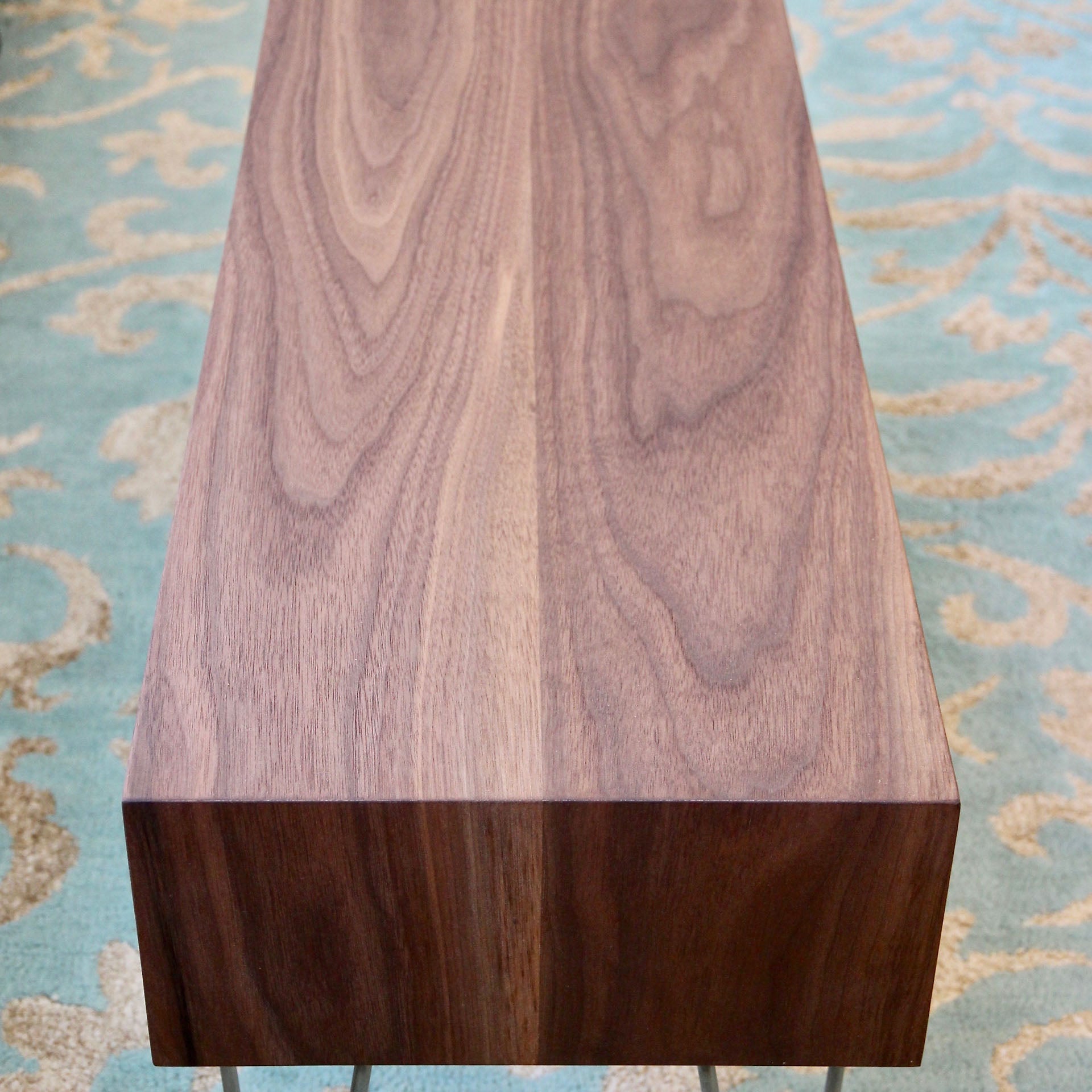 A substantial rectangular Coffee Table in Walnut, crafted by Krovel from Black Walnut and featuring a smooth surface with a natural grain pattern, rests on a blue and white patterned rug. The rich texture of the wood stands out beautifully against the ornate design of the carpet, capturing the essence of mid-century modern charm.