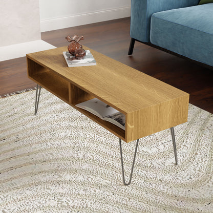 A modern living room showcases Krovel's Coffee Table in White Oak, complete with sleek stainless steel hairpin legs. The table holds a small decorative glass figure and a stack of magazines. It rests on a textured rug near a blue sofa, with the wooden flooring visible, exuding mid-century modern charm.