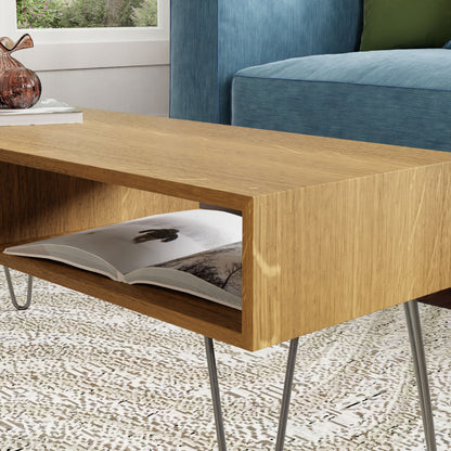 A White Oak coffee table from Krovel with stainless steel hairpin legs features a shelved area holding an open book. The table, embodying mid-century modern style, is placed on a patterned rug near a blue sofa with green and blue cushions. A decorative glass object adorns the tabletop.