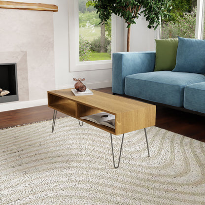 In a modern living room, a Coffee Table in White Oak by Krovel with stainless steel hairpin legs holds books and a decorative object. A textured rug covers the floor, and a blue sofa with green and gray cushions sits nearby. A fireplace and window provide views of lush greenery outside.