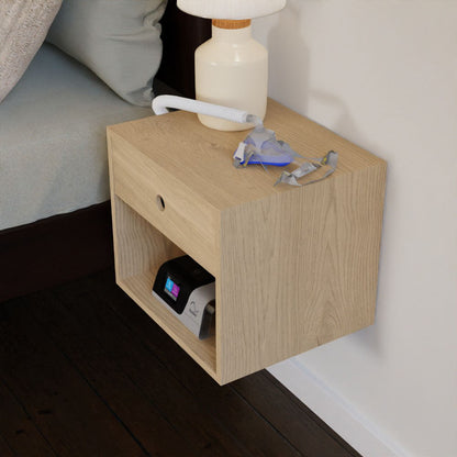 The CPAP Nightstand in Maple by Krovel exudes mid-century modern style with its solid maple construction and light wood grain finish. A table lamp and CPAP mask rest on the surface, while the open shelf below accommodates the CPAP machine. Placed next to a bed on a dark wooden floor, this nightstand artfully merges practicality with sophisticated design.