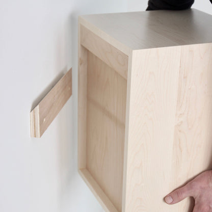 A person is mounting a Krovel CPAP Nightstand in Maple onto a wall using a wooden cleat for support. Reminiscent of Mid-Century Modern design, the solid maple nightstand is partially attached, with one piece of the cleat already fixed to the wall. The person is holding it in place similarly to a floating nightstand.