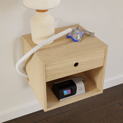 A CPAP Nightstand in Maple by Krovel, featuring a Mid-Century Modern design and crafted from solid maple, is mounted on the wall. It supports a lamp, CPAP mask, and hose on top. Below, the CPAP machine is placed on its open shelf against a white wall with dark hardwood floors beneath it.