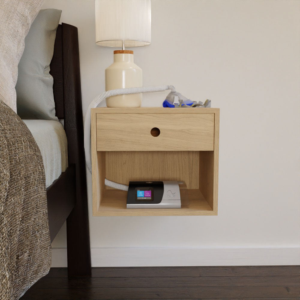 A CPAP Nightstand in Maple designed by Krovel showcases a CPAP machine neatly positioned on the lower shelf, with a beige lamp gracefully resting above. A hose is stylishly draped over the edge, while the bed partially enters the frame to the left, seamlessly integrating with the mid-century modern vibe of the hardwood floor beneath.