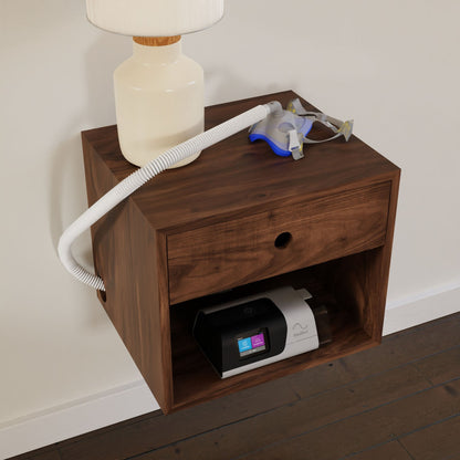 A CPAP Nightstand in Walnut from Krovel graces the white wall, embodying a Mid-Century Modern aesthetic. On top, a lamp and CPAP mask accompany the design. Below, a CPAP machine with a digital display rests on its shelf, set against the wooden floor.