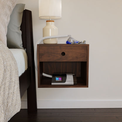 A CPAP machine from Krovel rests elegantly on a durable Walnut CPAP Nightstand, complete with an attached hose and mask. A beige lamp adorns the surface, exuding Mid-Century Modern charm. The bed sports a beige comforter and leans against a light-colored wall, enhancing the tranquil ambiance.