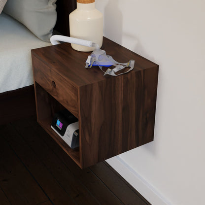 The Krovel CPAP Nightstand in Walnut, designed in a mid-century modern style and crafted from solid wood, is elegantly attached to the wall. It holds a CPAP machine with a nasal mask and hose on top, alongside a cream-colored bottle. The nightstand features a single drawer and an open shelf beneath it.