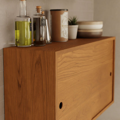 The Krovel Cupboard with Shelf in Cherry elegantly displays bottles of oil and vinegar, a small potted plant, and stacked bowls. This sturdy floating storage cabinet boasts a sliding door panel and is mounted on a neutral-toned wall using a French cleat, keeping items neatly organized on top.