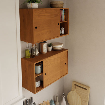 The Cupboard with Shelf in Cherry by Krovel is securely mounted on the wall with sliding doors. The upper section houses books, a plant, and dishes, while the lower section displays jars and bowls. Below, kitchen utensils and cutting boards are neatly arranged on a white tiled backsplash, all supported by a French cleat system.