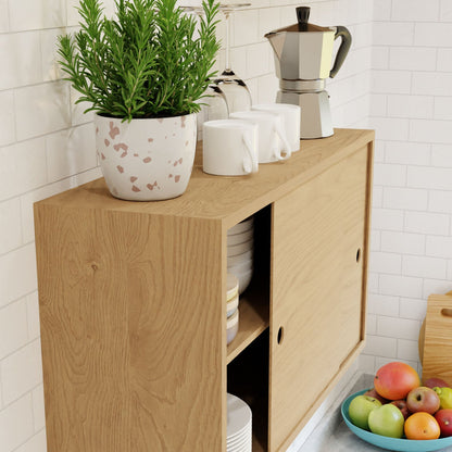 The "Cupboard with Shelf in Maple" by Krovel features white sliding doors and is set up as a floating storage cabinet for dishes. On top, it has a potted plant, three white mugs, a metal coffee maker, and glass holders mounted on the wall. Below it, there’s a counter with a bowl of fruit.