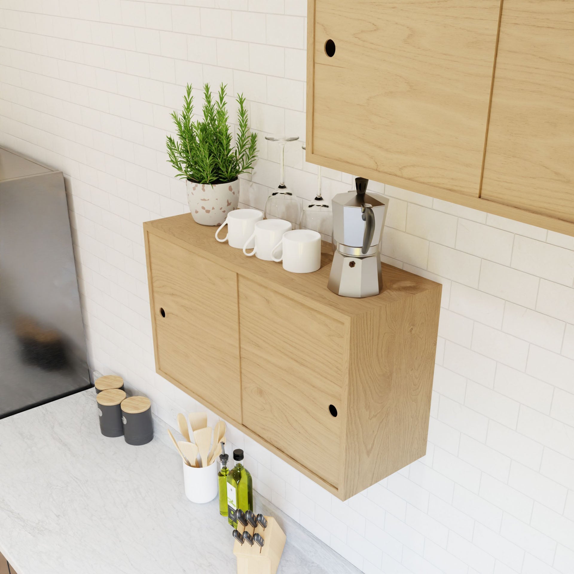 In a minimalist kitchen, light wood cabinets from Krovel's Cupboard with Shelf in Maple collection highlight floating storage mounted with a French cleat system. The countertop hosts spice jars, a wooden knife block, and olive oil bottles. A shelf is adorned with a plant, a moka pot, and three white mugs against the white tile backsplash.