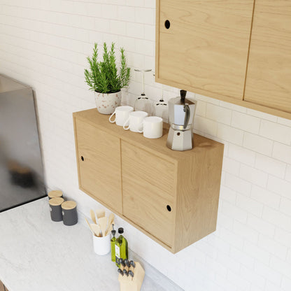 In a minimalist kitchen, light wood cabinets from Krovel's Cupboard with Shelf in Maple collection highlight floating storage mounted with a French cleat system. The countertop hosts spice jars, a wooden knife block, and olive oil bottles. A shelf is adorned with a plant, a moka pot, and three white mugs against the white tile backsplash.