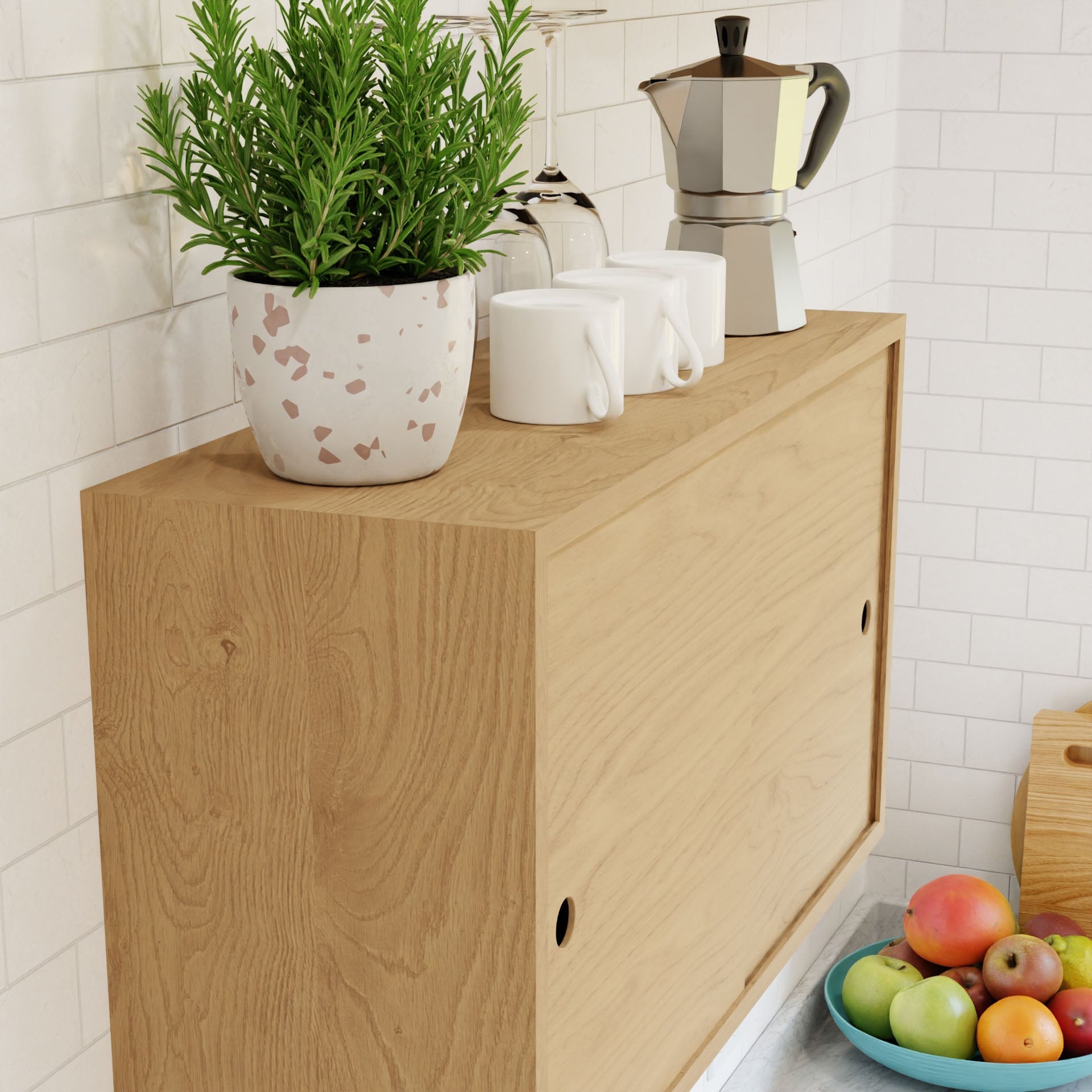 A maple cupboard with a shelf from Krovel in the kitchen holds three white mugs, a coffee maker, and a potted plant. Nearby, a white bowl contains apples and oranges. The background features white tiled walls, highlighting the Floating Storage Cabinet with its elegant French cleat design.