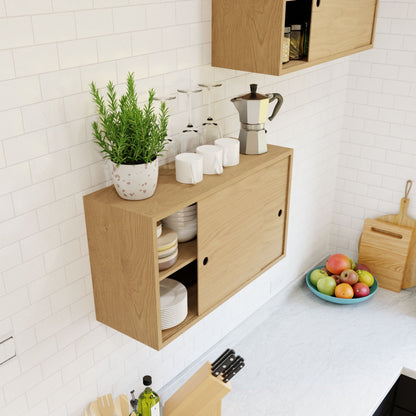 The "Cupboard with Shelf in Maple" by Krovel adds a rustic charm to the kitchen, holding white dishes and glasses behind its sliding door. A potted rosemary plant and a coffee maker are positioned on top beside a set of white mugs. Below, the solid walnut countertop displays a fruit bowl, knife set, cutting boards, and olive oil.