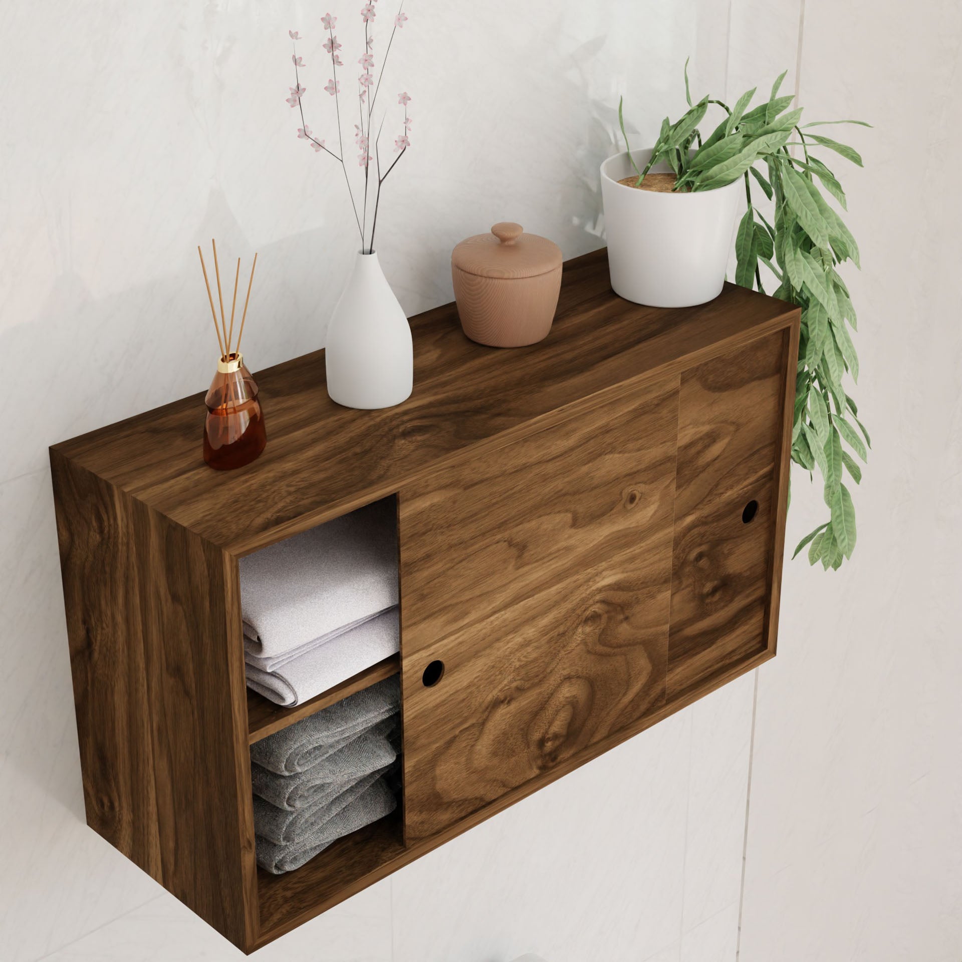 A Krovel Cupboard with Shelf in Walnut, featuring a wooden design and hand-finished for elegance, is mounted on the wall. It has sliding doors that reveal neatly folded towels on one side, while its top surface displays decorative items such as a vase with pink flowers, a small brown jar, a white pot housing green leaves, and a reed diffuser. This solid piece exudes both elegance and warmth.