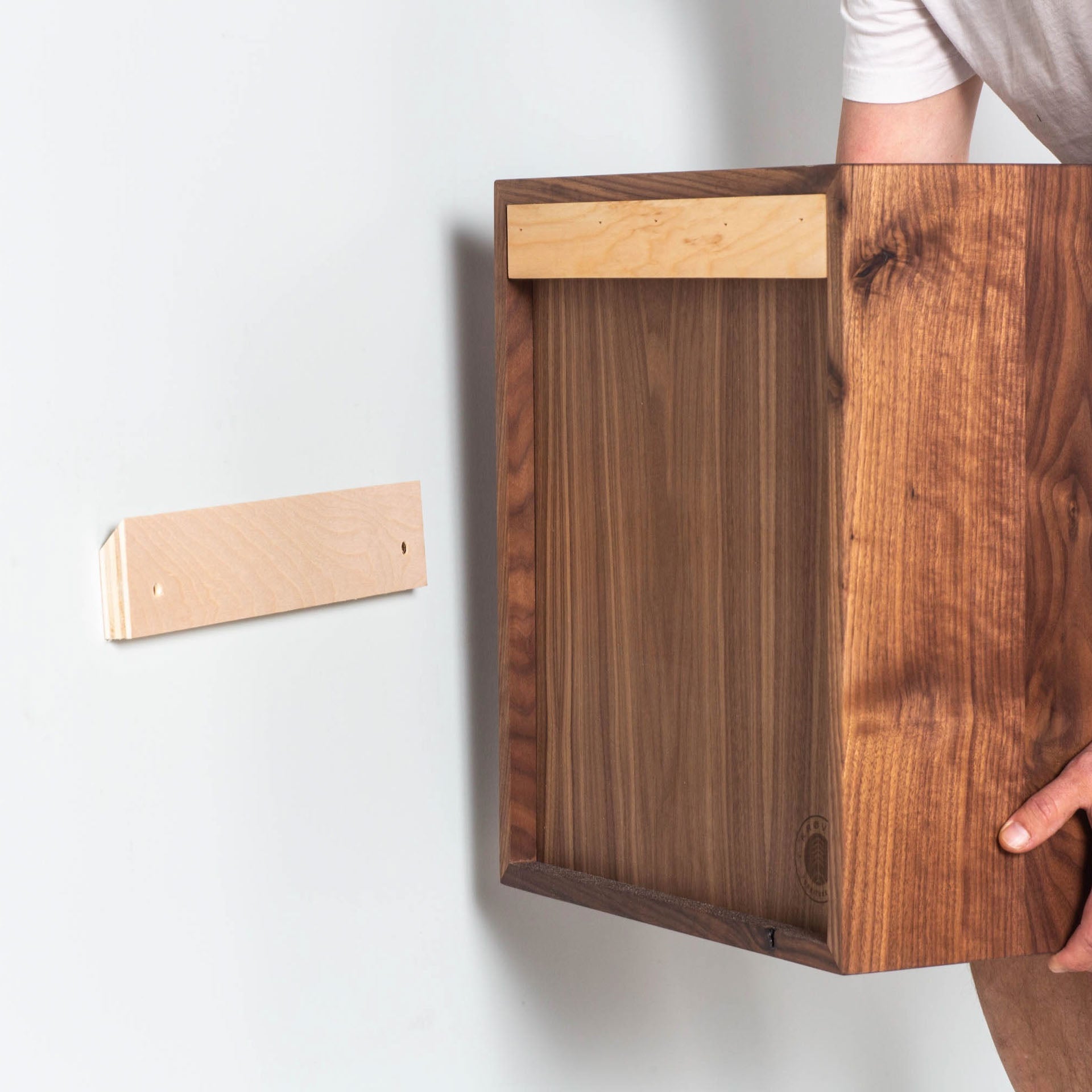 An individual is installing the Krovel Cupboard with Shelf in Walnut on a wall. The hand-finished wooden cabinet is being aligned with a bracket securely fastened to the wall, creating an elegant floating storage solution.