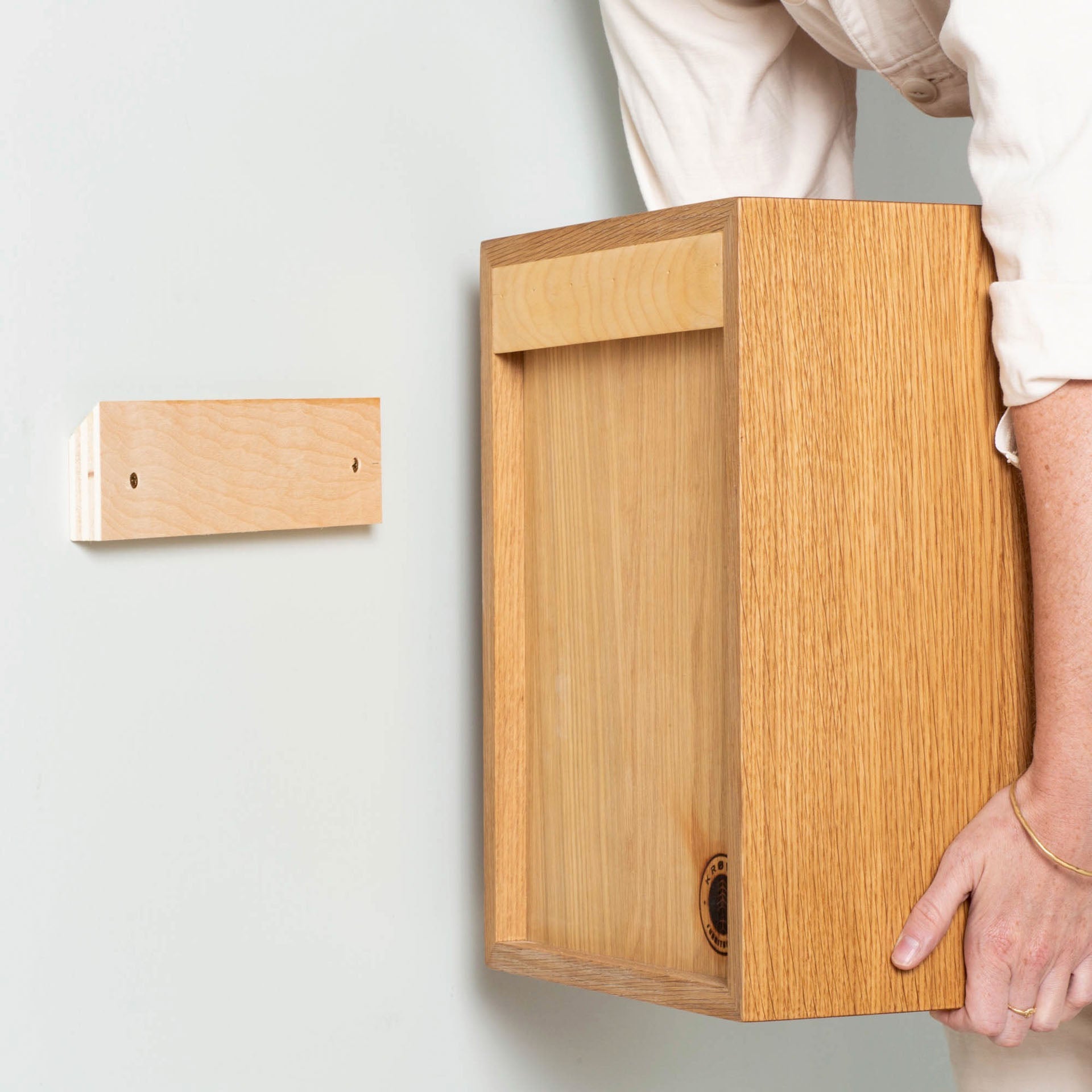 A person in a white shirt carefully mounts the Krovel Cupboard with Shelf in White Oak onto the wall using a wooden bracket. This floating storage cabinet features a light wood grain finish, adding charm and practicality to any space.