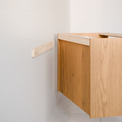 The Double Door Vanity in Cherry by Krovel Furniture Co. is being mounted on a pale wall with a wooden support rail. This floating vanity with hardwood veneer boasts a light brown wood finish, contrasting elegantly against the tiled white floor.