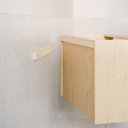 The Double Door Vanity in Maple by Krovel Furniture Co. features a minimalist design with hardwood veneer, wall-mounted with a light wood bracket, and placed against white marble tile walls. Its sleek aesthetic is enhanced by soft-close hinges for a modern touch.