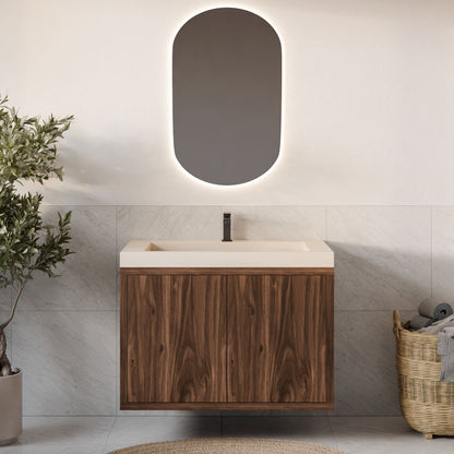 A modern bathroom features Krovel Furniture Co.'s Double Door Vanity in Walnut beneath an oval backlit mirror. Light gray tiles adorn the wall, complemented by a woven basket with towels on the right and a small olive tree in a pot on the left. A beige circular rug adds elegance to the floor.