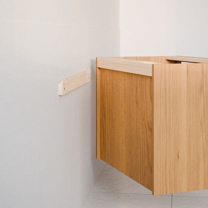 The Double Drawer, Open Shelf Vanity in Cherry by Krovel Furniture Co. is a floating wooden cabinet with a hardwood veneer, partially mounted to a white wall. Its cubic shape and natural wood finish include support from a wooden bracket, while the light floor below enhances its elegance.